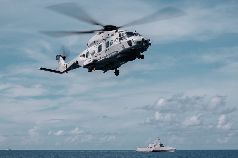 NH90 boven zee met op de achtergrond een marineschip.