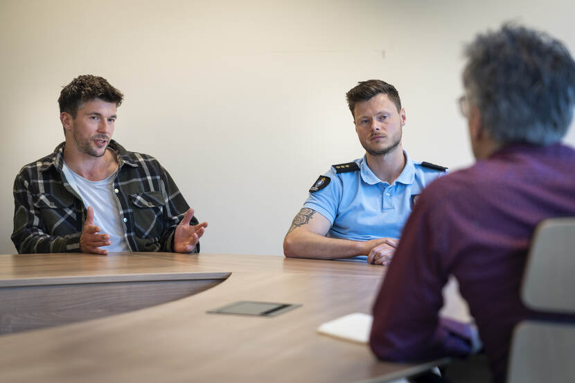 Man in geruit hemd en militair in blauw shirt zitten aan kantoortafel tijdens interview.