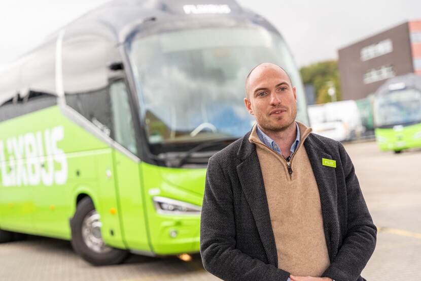 Portret van man met bruine trui en zwart jasje die voor een groene autobus staat.