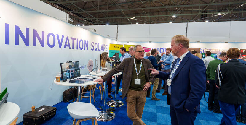 Twee beursbezoekers in gesprek bij een statafel naast een lange muur met de tekst Innovation Square.