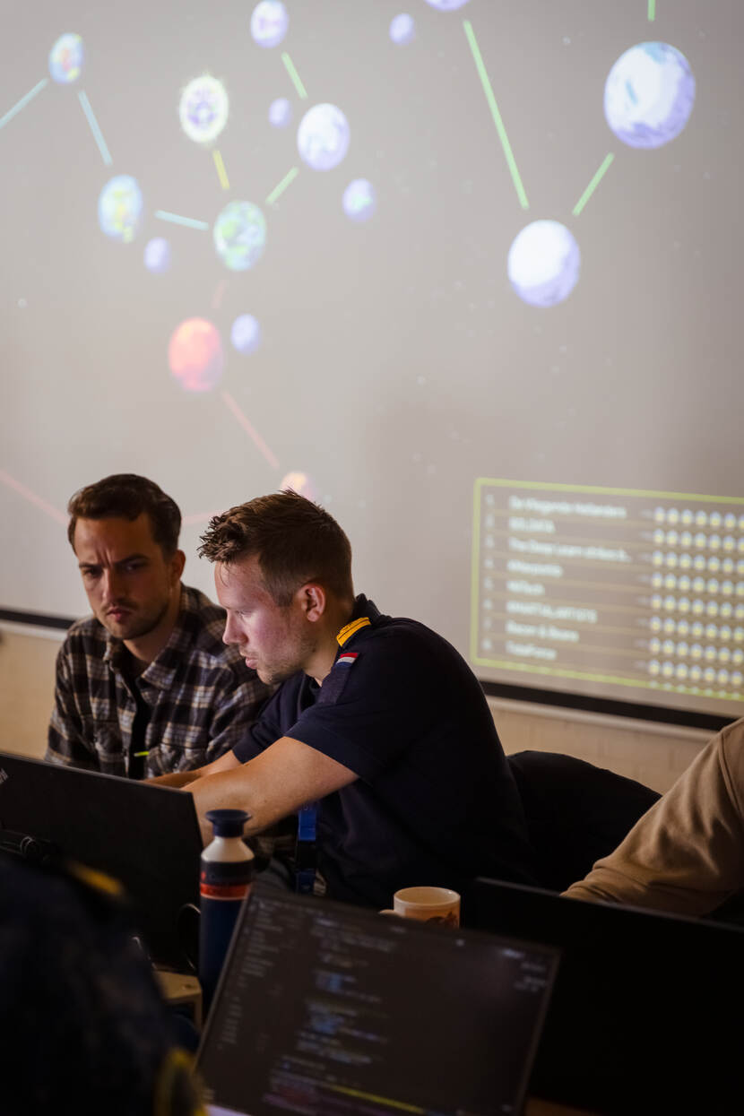 2 personen achter een laptop met op de achtergrond groot videoscherm.