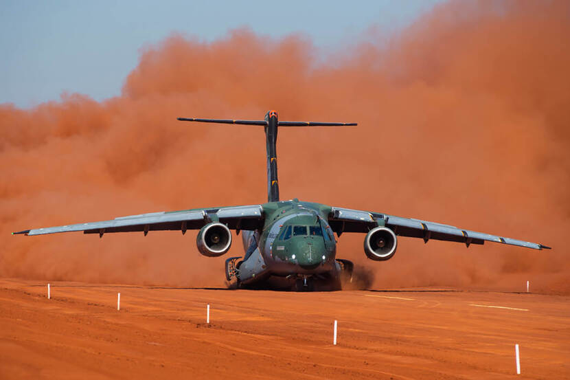 Een Embraer C-390 landt op een zanderige vlakte. Aan weerszijden stijgen stofwolken op.