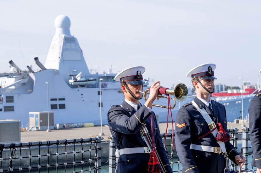Twee mannen in net pak van de marine. Een van hen blaast op een trompet. Op de achtergrond ligt het nieuwe CSS Den Helder.