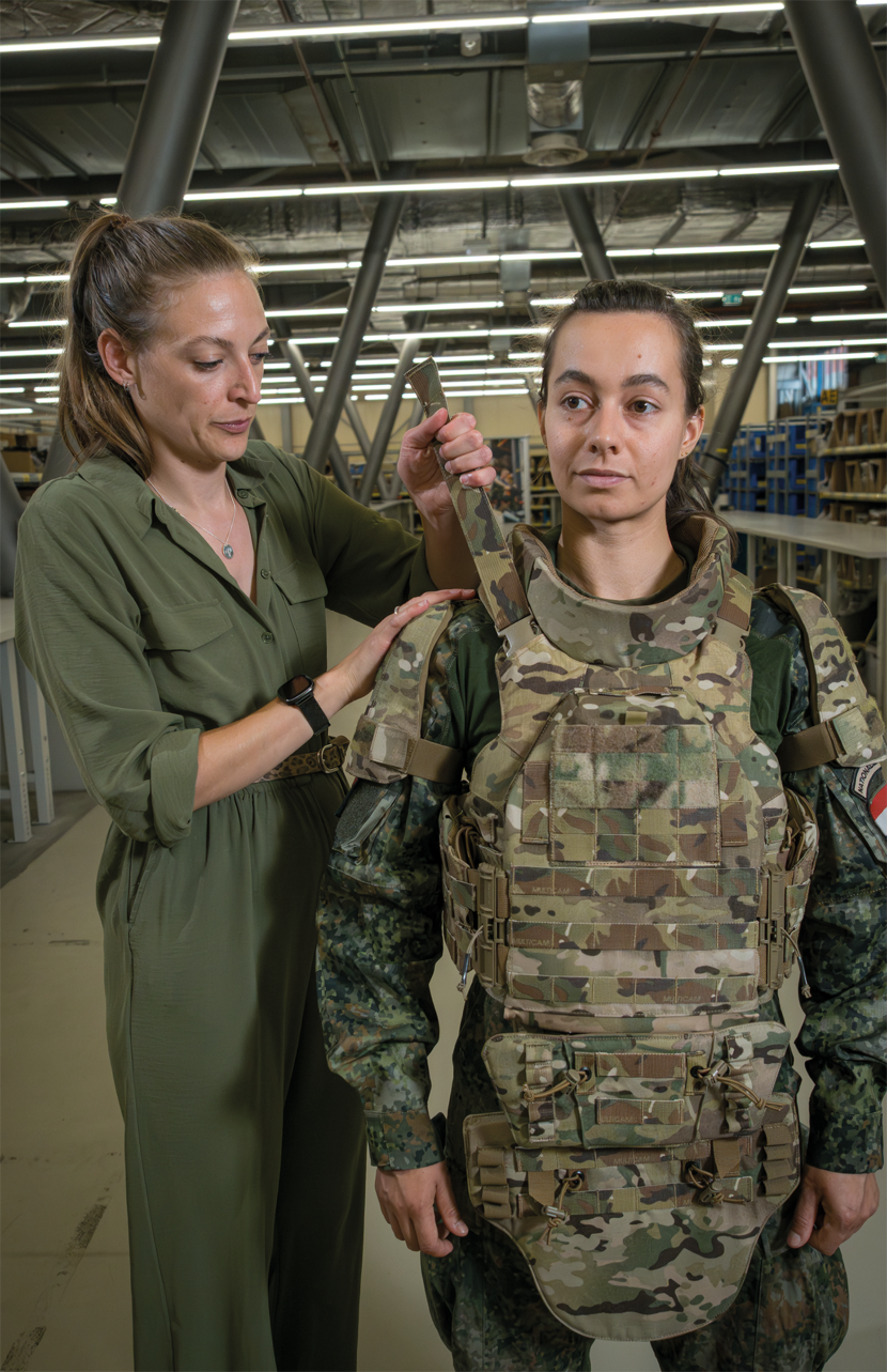 Een vrouwelijke militair draagt het scherfvest voor vrouwen. Schuin achter haar maakt een medewerker van het KPU-bedrijf het vest op maat.