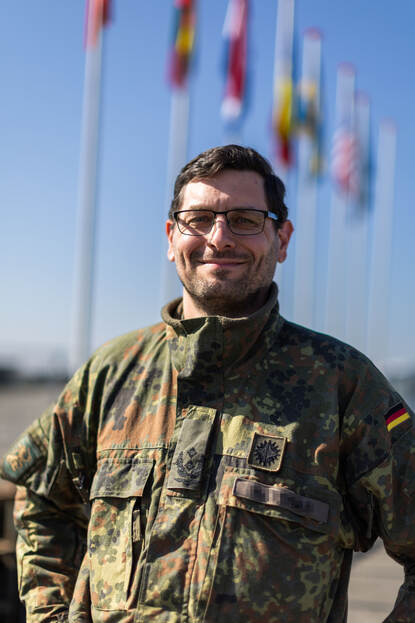 Portretfoto van luitenant-kolonel Benjamin. Achter hem zijn de vlaggen van de deelnemende landen te zien.