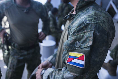 Links bewaakt een marinier een gevangengenomen vijand. Rechts een mouwembleem waarbij de Nederlandse en Colombiaanse vlag in elkaar overlopen.