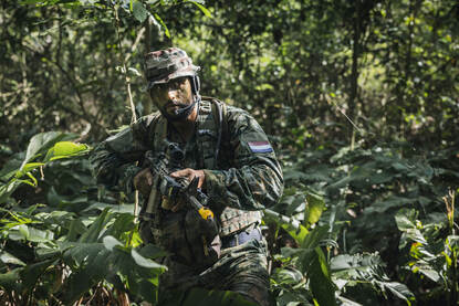 Links sluipt een marinier volledig gecamoufleerd door de jungle. Rechts een hagedis met een rode kop op een boomstam.