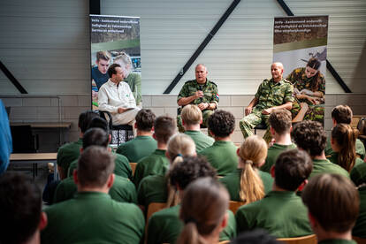 Links: Studenten zitten op een rij naast elkaar. Een van hen leest een vraag op van een briefje. Rechts: Twee militairen zitten met presentator op podium in zaal vol studenten.