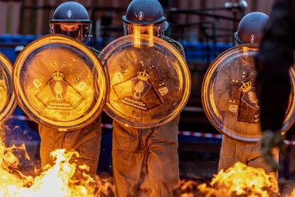 Links; Marechaussee in camouflagepak marcheren. Rechts; Marechaussees met helm en schild lopen door vuur in ME-tenue.