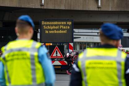 Links een protestbord met de tekst ‘Deze weg loopt dood’, rechts silhouetten van twee Marechaussees met op de achtergrond een matrixbord met de tekst ‘Demonstreren op Schiphol Plaza is verboden’.