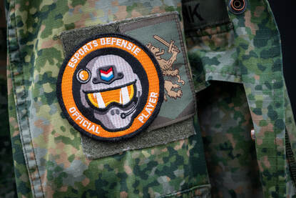 Embleem op schouder militair van helm met oranje vizier en de Nederlandse vlag op het voorhoofd. Rondom staat de tekst ‘Esports Defensie Official Player’.