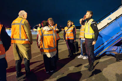 Vrouw in oranje hesje wordt onderaan vliegtuigtrap welkom geheten terwijl Marechaussee salueert.