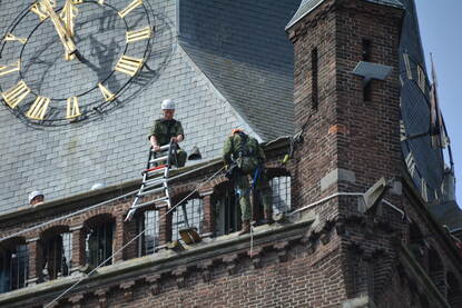 Militairen dalen met een touw af van de kerk, rechts twee lachende vrouwen die veel plezier beleven aan het evenement.