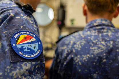 Boven het logo van Sandy Coast 2025 op de mouw van een marineman en duikondersteuningsvaartuig Zr.Ms. Nautilus aan de kade van Eemshaven. De DSS Galatea op zee en een rubber boot die over het water scheert.