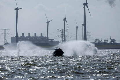 Boven het logo van Sandy Coast 2025 op de mouw van een marineman en duikondersteuningsvaartuig Zr.Ms. Nautilus aan de kade van Eemshaven. De DSS Galatea op zee en een rubber boot die over het water scheert.