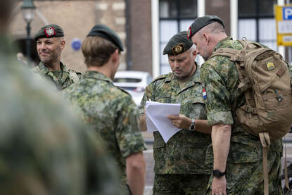 Een groepje militairen loopt samen tussen de burgers. De voorste militair heeft een plattegrond in zijn handen. Achter hen staat een bus geparkeerd. Daarnaast vier militairen die in overleg zijn met elkaar.