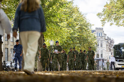 Een groepje militairen loopt samen tussen de burgers. De voorste militair heeft een plattegrond in zijn handen. Achter hen staat een bus geparkeerd. Daarnaast vier militairen die in overleg zijn met elkaar.