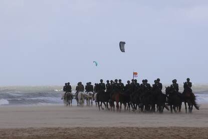 links: een fanfare speelt op het strand, op de achtergrond ruiters te paard. Rechts: Paarden rennen over het strand terwijl zand opstuift.