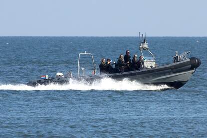 Links een grijze militaire helikopter met draaiende rotors en ingetrokken landinggestel vliegt tegen een helderblauwe lucht. Rechts een snelle, grijze militaire speedboot met vijf bemanningsleden vaart over rustig, blauw zeewater met witte golven achterop.