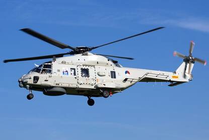 Links een grijze militaire helikopter met draaiende rotors en ingetrokken landinggestel vliegt tegen een helderblauwe lucht. Rechts een snelle, grijze militaire speedboot met vijf bemanningsleden vaart over rustig, blauw zeewater met witte golven achterop.