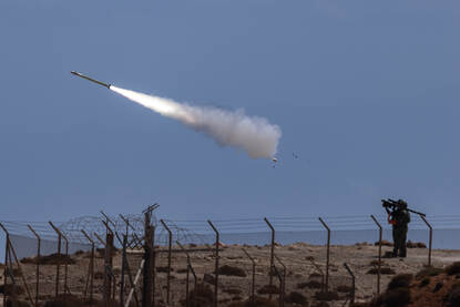 Links: Een raket wordt afgeschoten vanaf militair voertuig met veel rook en vuur, tegen een heuvelachtige, droge en rotsige achtergrond. Rechts: een soldaat die een raket afvuurt vanaf een heuvel met prikkeldraad, terwijl de raket met witte rooksporen door de lucht schiet.