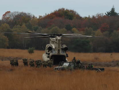 Links een helikopter vliegt in het schemer door de lucht met een voertuig aan een touw er onder. Rechts: Militairen stappen in een helikopter op de heidegrond.