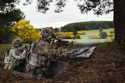 twee militairen in camouflagetenue liggen achter een lage muur, gericht op de verte met een groot geweer op een statief, omringd door bos en open veld. Rechts: Twee soldaten in camouflagetenue staan in een bos; één helpt de ander met zijn rugzak, terwijl de ander een geweer vasthoudt en iets roept.