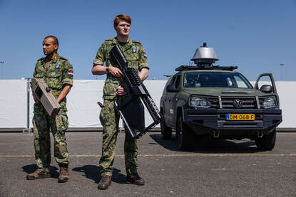 Links in beeld een NASAMS-lanceerinstallatie opgesteld op een parkeerplaats in de duinen. Rechts twee militairen met anti-drone-middelen.