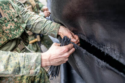 Links een close-up van handen die tie-wraps bevestigen. Rechts twee militairen die een zwart zeildoek spannen.