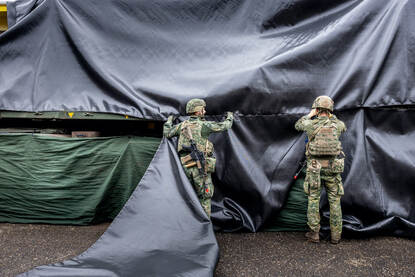 Links een close-up van handen die tie-wraps bevestigen. Rechts twee militairen die een zwart zeildoek spannen.