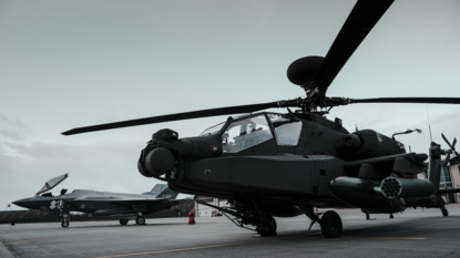 Apache met draaiende rotorbladen staat op een militaire landingsbaan; op de achtergrond is een F-35 geparkeerd onder een bewolkte lucht.