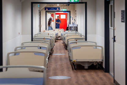 Links rijen oude ziekenhuisbedden op een gang van het ziekenhuis. Rechts de bedden opgesteld in een lange vrachtwagenaanhanger.