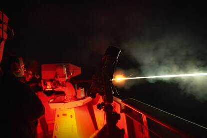 Links schiet een wapen met een lichtspoor in het donker. Rechts varen twee fregatten in de schemering langs een boorplatform.