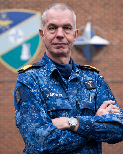CDR Arjen Warnaar met op de achtergrond het NAVO-embleem en het van het Allied Maritime Command.