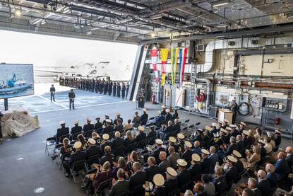 Genodigden zitten op stoelen op marineschip tijdens ceremonie en de muzikale begeleiding tijdens ceremonie.