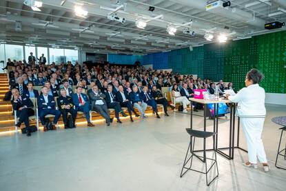 Vrouw in wit pak spreekt volle zaal toe.