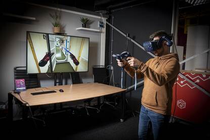 Op de foto staat een man met VR-bril op, die met in elke hand een controller voor videoscherm staat waarop de binnenkant van een marineschip virtueel is weergegeven.