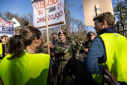Militairen gaan voor ingang militair terrein in gesprek met groepje demonstranten met spandoeken en hesjes aan.