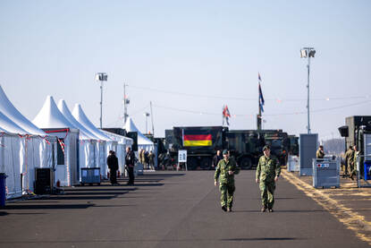 Over het oefenterrein lopen temidden van witte tenten 2 landmachtmilitairen.