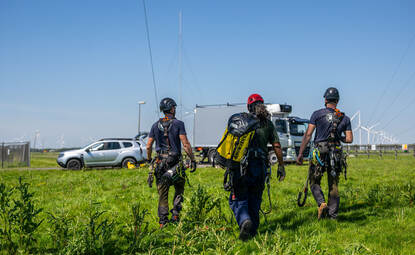 Drie technici op de rug gezien met klimuitrusting lopen veld in.