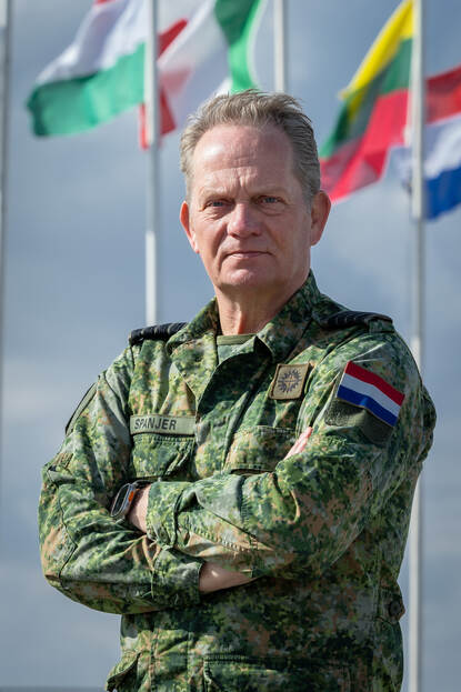 Portretfoto van kolonel Olav Spanjer. Op de achtergrond wapperen vlaggen van onder meer Italië, Litouwen en Nederland.