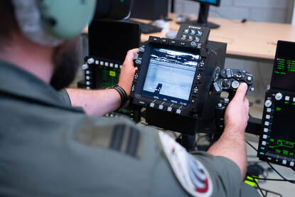 Een vlieger zit in een bureaustoel met een VR-bril op zij hoofd. Ook draagt hij een koptelefoon.