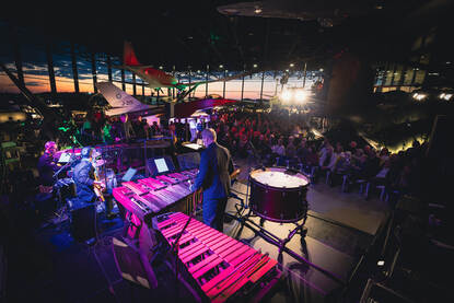 Slagwerkers van het Orkest Koninklijke Luchtmacht met op de achtergrond een aantal vliegtuigen van het museum en een ondergaande zon.