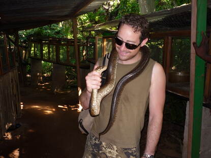 Foto links: Martijn tuurt vanaf een schip door zijn verrekijker. Foto rechts: Martijn met een slang om zijn nek in een tropisch bos.