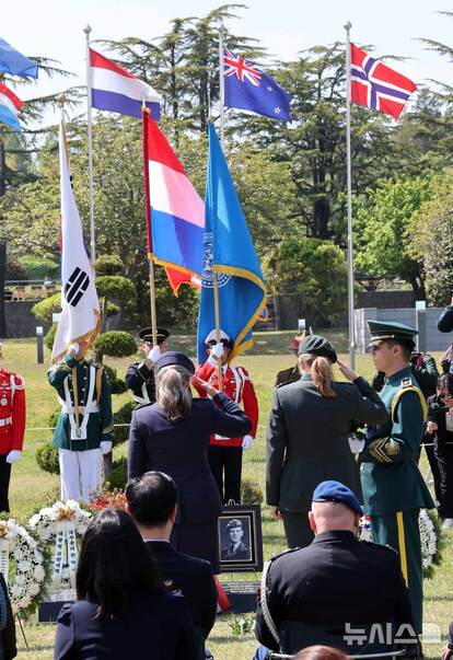 Twee Nederlandse vrouwelijke militairen brengen militairen groet op een begraafplaats aan een foto van Nederlandse militair. Op de achtergrond diverse landsvlaggen.