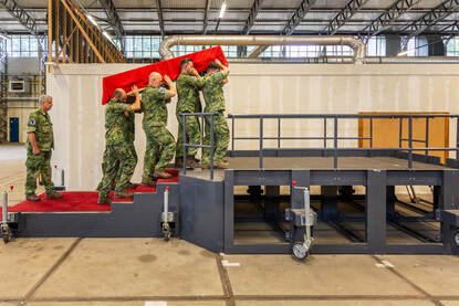 De draagploeg loopt met de lijkkist op een replica van de trap bij Paleis Noordeinde. Daarop ligt een rode loper. Daarnaast een viertal dat zich, met de armen over elkaar heen geslagen, naar boven verplaatst.’