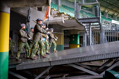 De vaandelwacht van de luchtmacht betreedt het veld vanuit een hoek van het stadion. De vaandeldrager in het midden, daaromheen militairen die een Colt C8-geweer vasthouden.