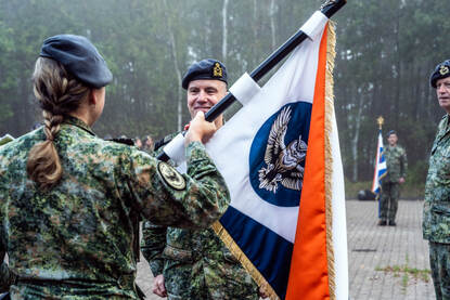 Een militair met een vlag in zijn handen staat tegenover een militair die de vlag in ontvangst neemt.