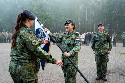 Een militair met een vlag in haar handen staat tegenover een militair die de vlag in ontvangst gaat nemen. Een derde militair kijkt aan de zijkant toe.