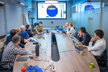 Mensen aan een lange ovale vergadertafel met een groot beeldscherm op de kopse kant.